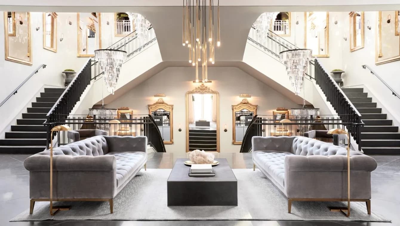 This is an image from J & S Painting Plus, located in New York. The interior photo highlights a grand living area with twin staircases, tufted gray sofas, chandeliers, and symmetrical architectural details.