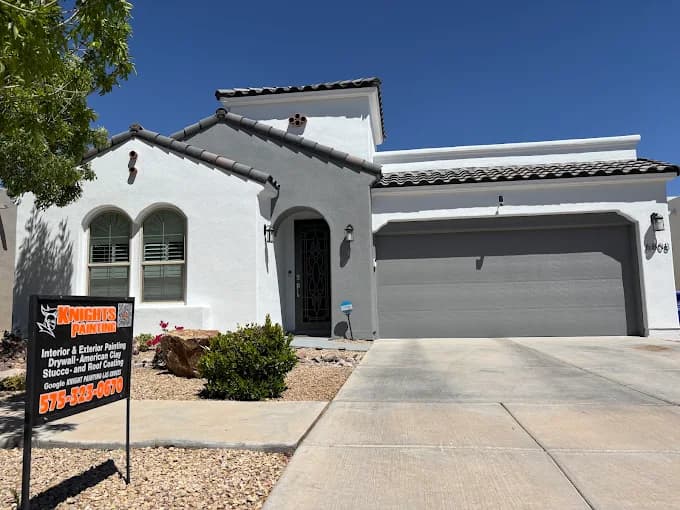 This is an image from Knight's Painting, located in New Mexico. The photo shows a newly painted modern stucco home with white walls, gray trim, and a gray garage door, with a company yard sign displayed in the front yard.