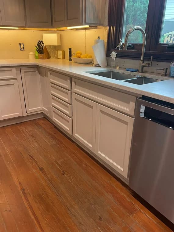 This is an image from Ponderosa Painting LLC, located in Louisiana. The photo shows a freshly painted kitchen with light gray cabinetry, marble countertops, and natural wood flooring, emphasizing detailed interior finish work around the sink and window area.