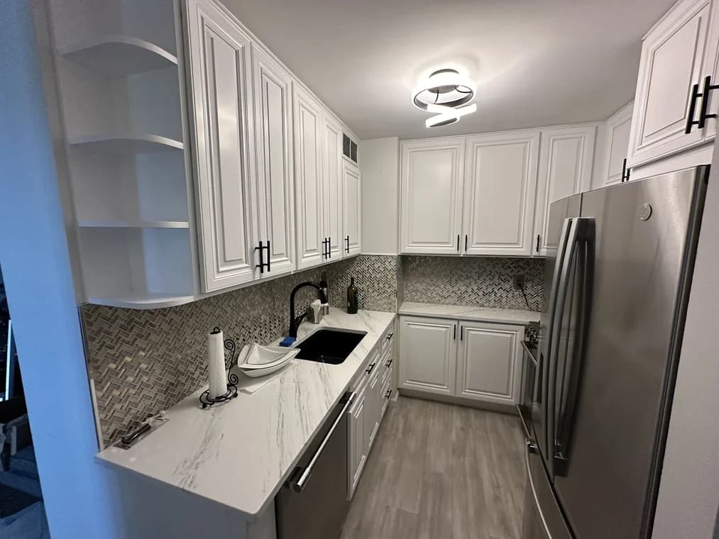 This is an image from Nyca Contractors, LLC, located in New York. The kitchen features freshly installed white cabinets with black hardware, a patterned backsplash, stainless steel appliances, and a modern circular ceiling light.