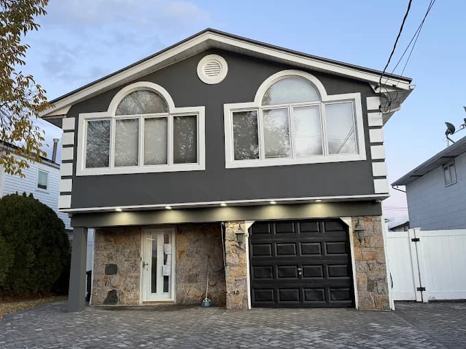 This is an image from Premier Painting Pros, located in New York. The photo shows a two-story home with a dark gray exterior, white trim, arched windows, and stonework around the entry and garage.