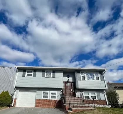 This is an image from By Hand Painting LLC, located in New Jersey. The photo shows a two-story gray home with white trim and a brick foundation, viewed from the driveway under a partly cloudy blue sky.