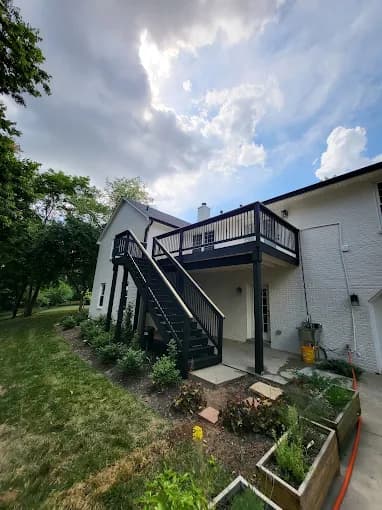 This is an image from Skywalkers Painting & Repairs, located in Ohio. The image shows a white brick home exterior with a newly painted elevated deck and black staircase overlooking a landscaped backyard.