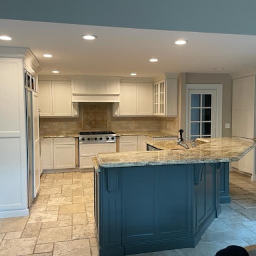 This is an image from Big Expert Painting LLC, located in New York. The kitchen features freshly painted white cabinets, a contrasting blue island, recessed lighting, and stone tile flooring for a clean updated look.