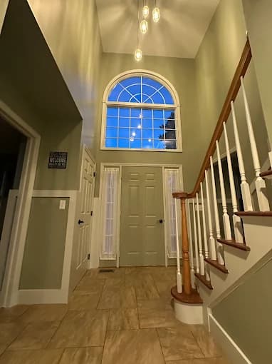This is an image from Richard K. Moore Professional Painting, located in New Jersey. The image shows a freshly painted residential entryway with soft green walls, white trim, a staircase with white balusters and wood handrail, and a tall arched window bringing in natural light.