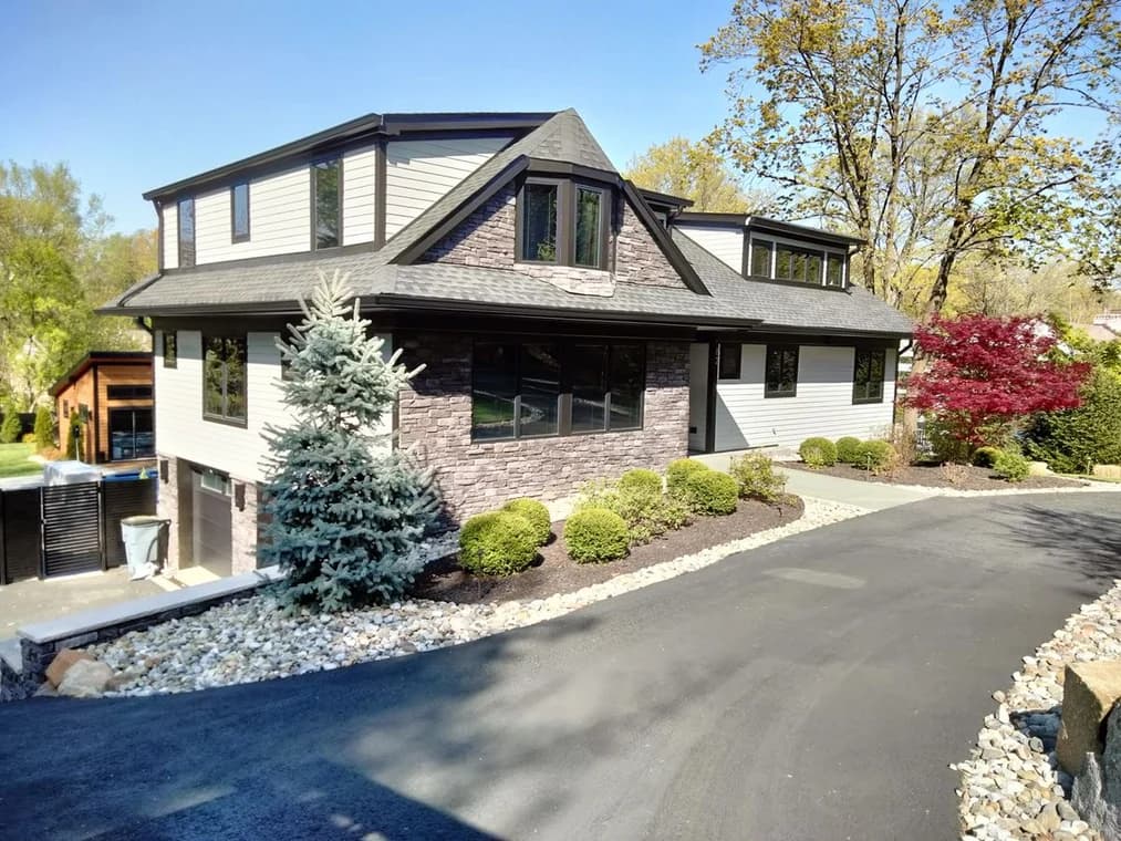 A modern renovated home with light siding, stone accents, dark trim, and landscaped front yard with shrubs, trees, and a paved driveway.