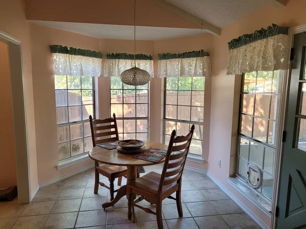 "This is an image from Hutch-N-Son Painting & Drywall, located in Texas. The photo shows a breakfast nook with peach-colored walls, tile flooring, and multiple tall windows with lace valances."