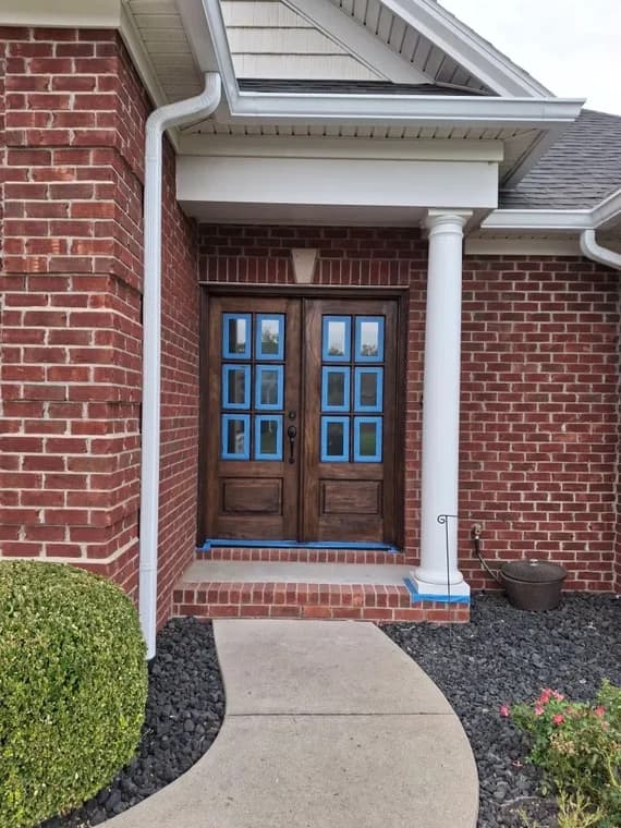 This is an image from Triple G Custom Painting Drywall & Roofing LLC, located in Indiana. The photo shows a front entryway with double wooden doors on a red brick home, with blue painter’s tape outlining the glass panes and trim for a clean finish.