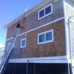 This is an image from Promark Painting, located in Massachusetts. It shows a worker on a tall ladder preparing a multi-level home’s exterior shingles for painting, demonstrating their restoration process.