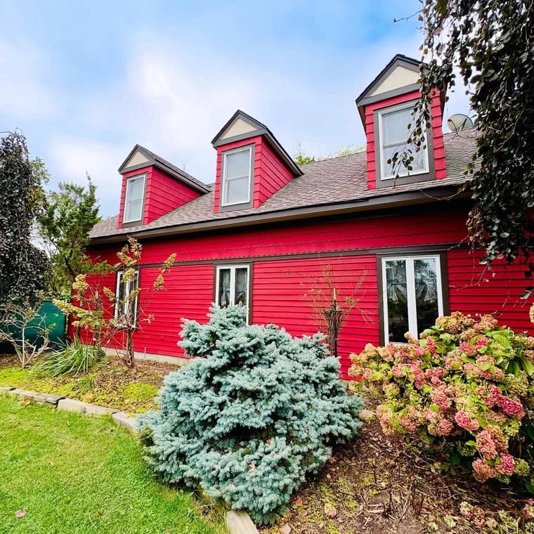 This is an image from Brush House Painting & Flooring Services of North Jersey, located in New Jersey. The image shows a freshly painted bright red home exterior with dormer windows, gray trim, and landscaped shrubs along the front yard.