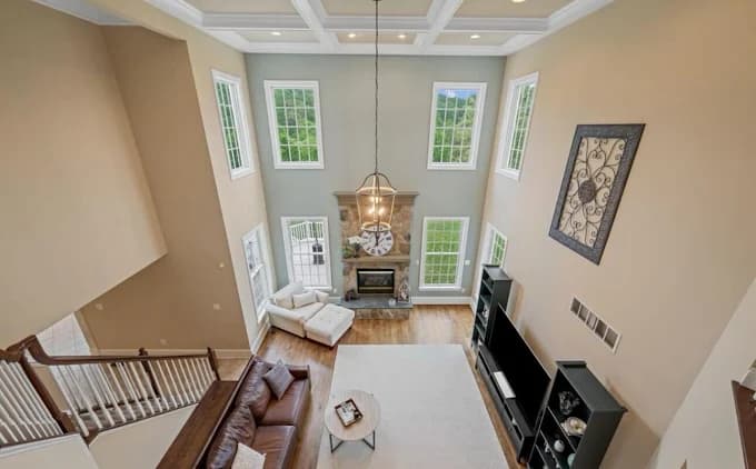 This is an image from Perfect Painters & More, located in Maryland. The photo shows a spacious living room with freshly painted neutral walls, tall windows, a coffered ceiling, and a stone fireplace as the centerpiece.