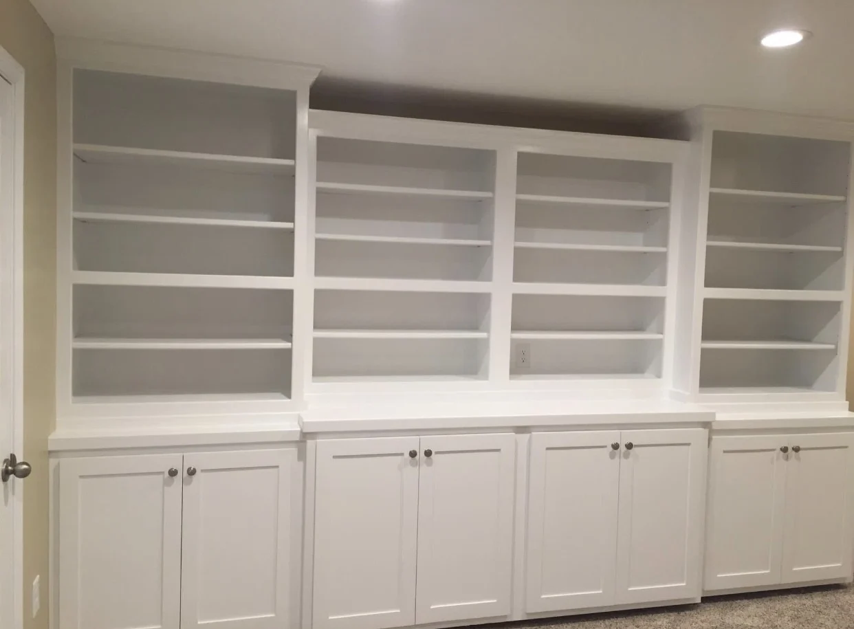 This is an image from Brogan Painting & Remodeling. The image shows freshly painted white built-in shelving with lower cabinets and clean, even finish work.