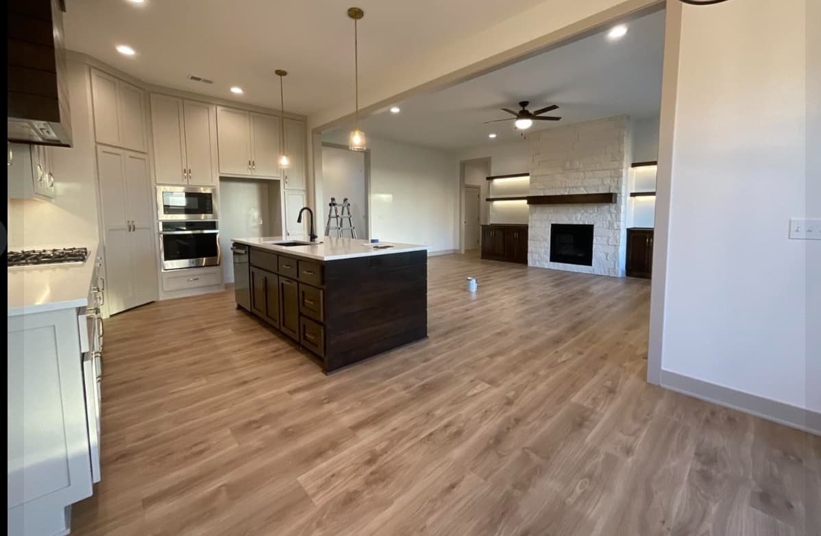 This is an image from Painter's Painting. The image is a Open kitchen and living room area in Kansas home with newly painted white walls, wood cabinets, and stone fireplace.