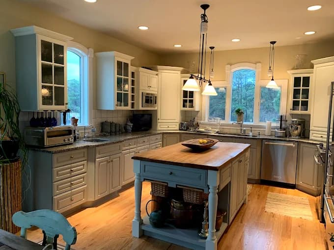 This is an image from Chameleon Painting Inc., located in New York. The photo shows a warm, inviting kitchen with cream-colored cabinets, granite countertops, stainless steel appliances, and a central island with a wooden top.