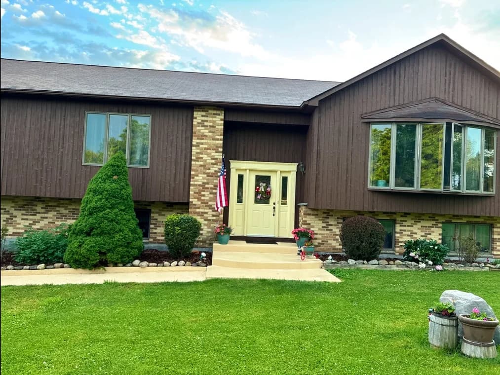 This is an image from Certified Painting, located in New York. The photo shows a two-story split-level home with dark brown vertical siding, brick accents, and a cream-colored front door surrounded by neatly maintained landscaping.
