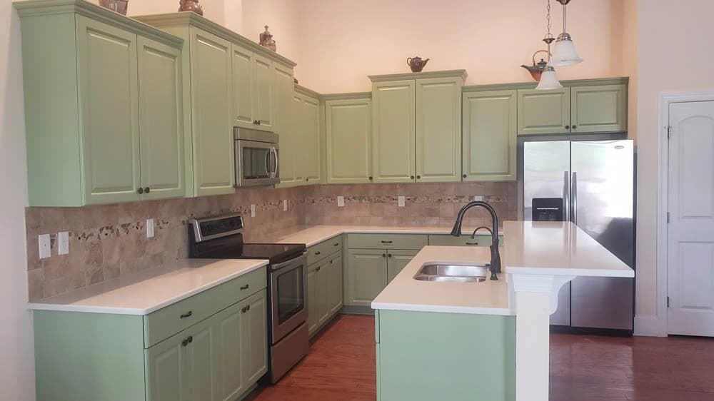 Cabinet painting project by R3 Painting in Hampstead, NC. The image shows a kitchen with pale green cabinets, tile backsplash, counters, and high ceiling.
