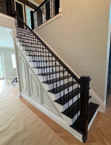 This is an image from Teall Painting, located in Michigan. The image shows a detailed interior staircase renovation with painted risers, dark handrails, black iron balusters, and crisp white wall paneling.