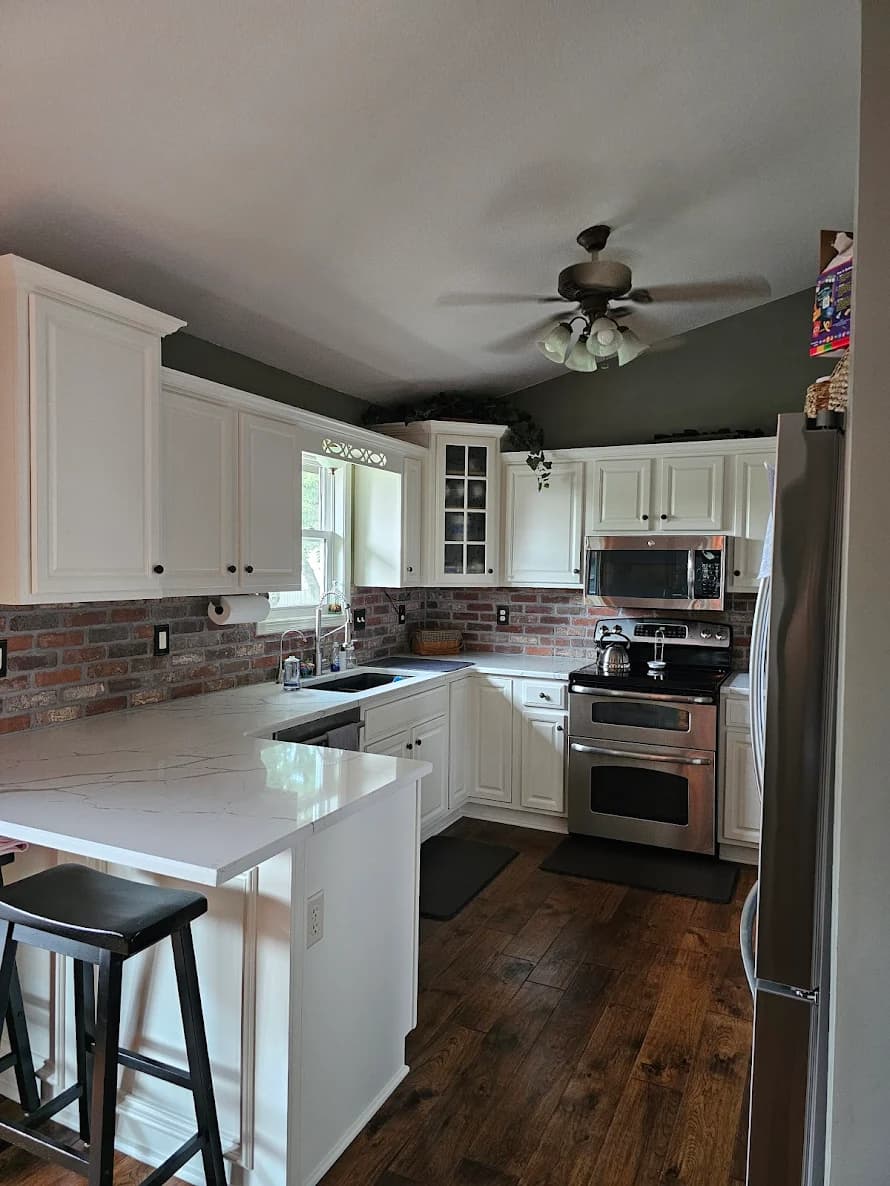 This is an image from River City Painting Inc. The image shows a fully repainted kitchen featuring crisp white cabinets, updated hardware, and a clean brick backsplash, highlighting the refreshed interior painting work.