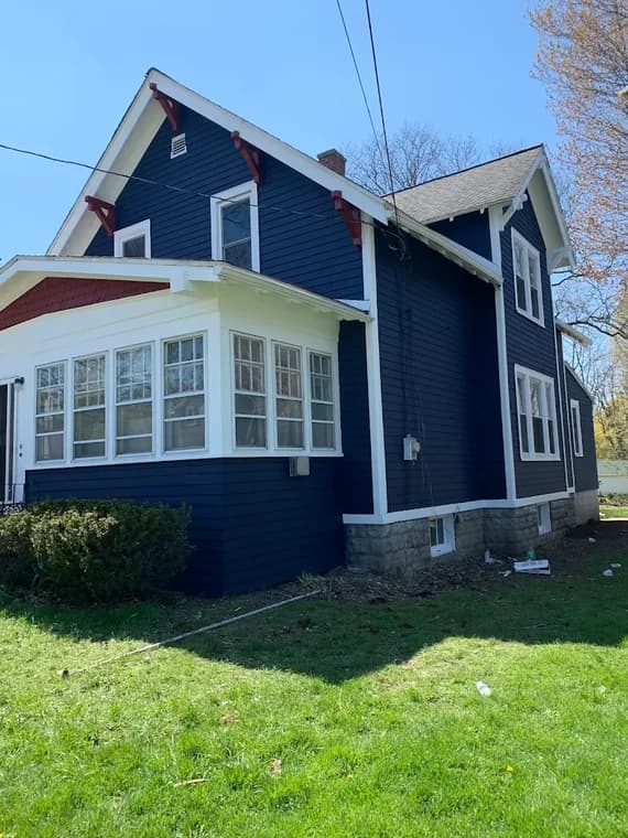 This is an image from Northeast Pro Painting & Restoration, located in New York. The picture shows a dark blue house with white trim and red accent brackets, showcasing a completed exterior repaint with crisp detailing around windows and rooflines.