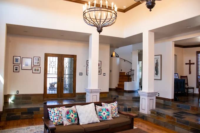 This is an image from Mike’s Quality Painting, located in New Mexico. The photo shows a beautifully painted interior living space with smooth white walls, warm wooden trim, slate flooring, and a chandelier above a cozy brown sofa with colorful patterned pillows.