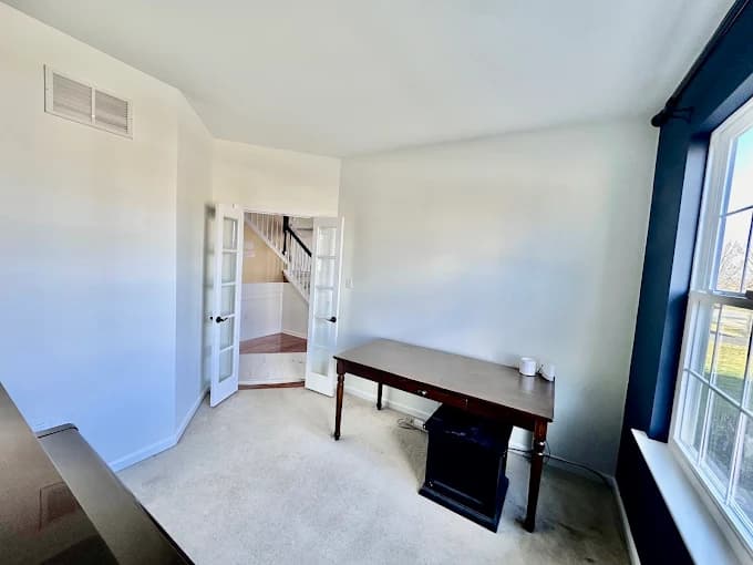 This is an image from Pro Painters Plus LLC company, located in Perkasie, PA. The image is a freshly painted small home office with white walls, navy accent wall by the window, beige carpet, a wooden desk, and double glass French doors leading to a hallway with staircase.