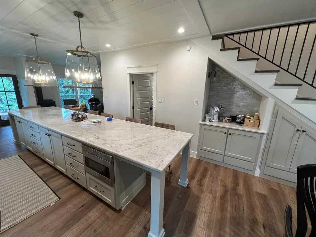 This is an image from Yellow Rose Painting, located in Texas. The photo shows a modern kitchen with a long marble island, light gray cabinetry, pendant lights, and warm wood flooring.