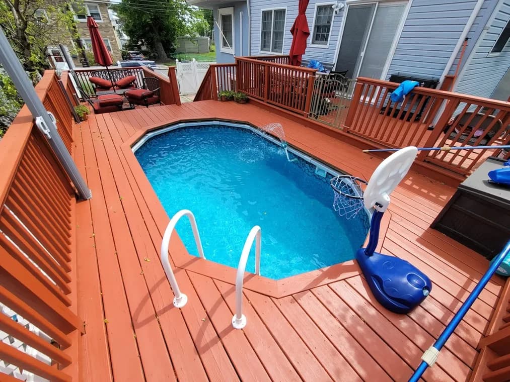 This is an image from CertaPro Painters of Staten Island, located in New York. The image shows a red-painted wooden deck surrounding an above-ground pool with clear blue water and lounge seating in the background.