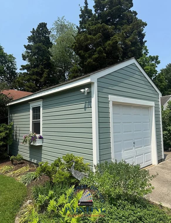 This is an image from Original Design Painting, showing a detached garage painted in a muted green tone with white trim. The garage sits among lush greenery and landscaped plants.