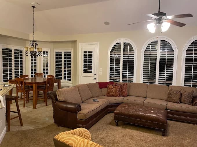 This is an image from Platinum Painting of Richardson, located in Texas. The photo shows a warm-toned living room with a large sectional sofa, ceiling fan, and freshly painted beige walls surrounding arched windows with white shutters.