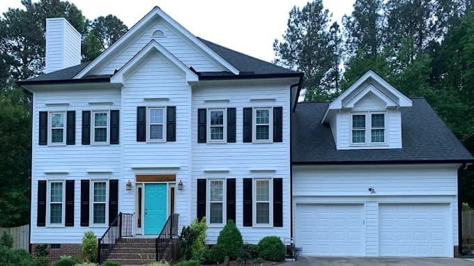 This is an image from Gonzalez Painters & Contractors, Inc., located in North Carolina. The photo shows a beautifully painted two-story white house with black shutters, a teal front door, and an attached garage surrounded by trees.