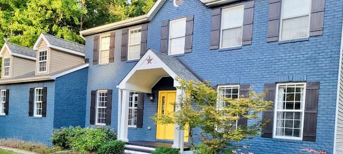 This is an image from Perfect Painters & More, located in Maryland. The photo shows a two-story brick home freshly painted in a deep blue color with white trim, brown shutters, and a bright yellow front door.