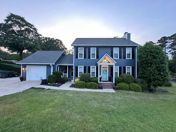 Exterior painting project by Garner's Painting Service in Fayetteville, North Carolina. The image shows a blue two-story home with white trim, red shutters, a garage, and lawn.
