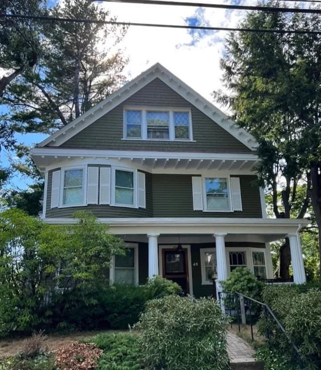 This is an image from Ulta Home, located in Massachusetts. It features a classic-style house with olive-green siding, white shutters, and a front porch surrounded by trees, reflecting the company’s exterior painting services.