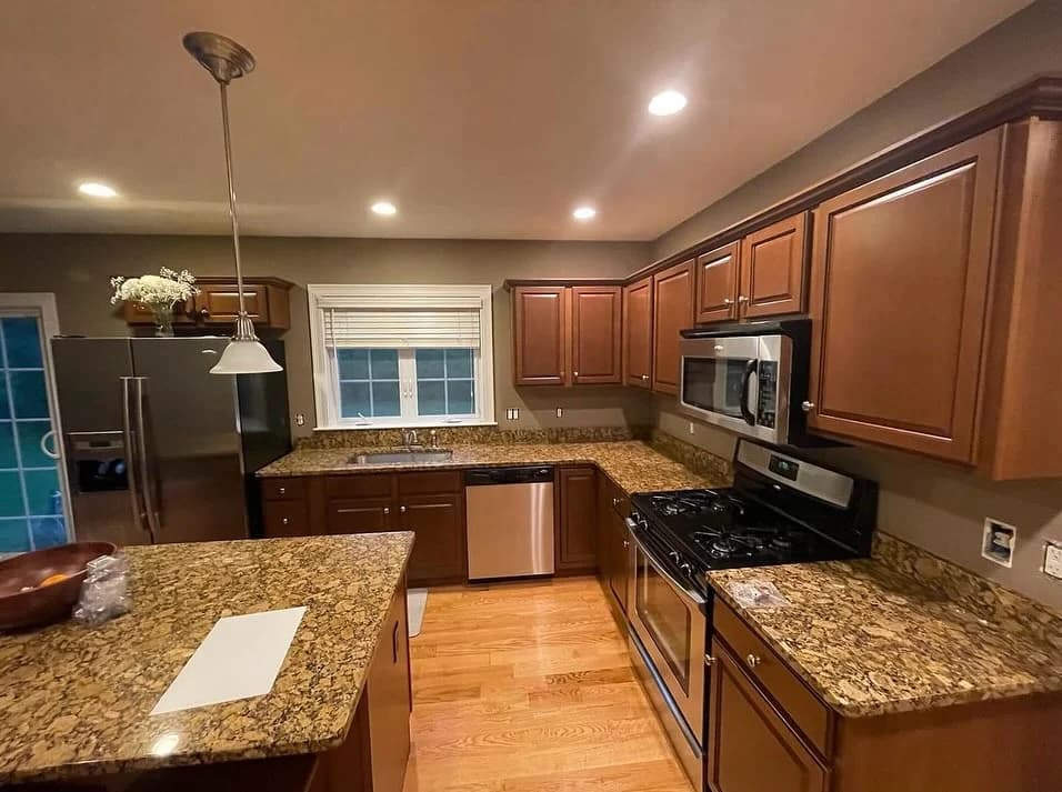 This is an image from EVM Home Improvement & Painting, located in Massachusetts. The photo shows a modern kitchen with granite countertops, wooden cabinetry, stainless steel appliances, and recessed lighting.