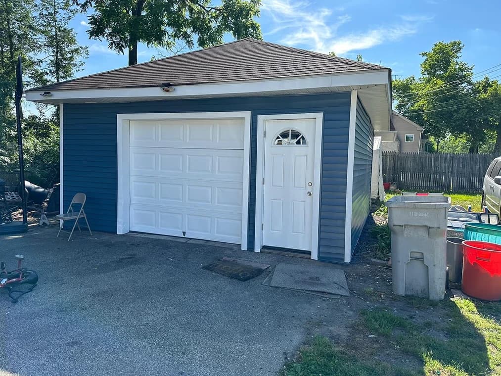 This is an image from Clean Lines Painting & Powerwashing of New Jersey, the image is a detached garage with dark blue siding and crisp white trim and doors, repainted by Clean Lines Painting + Powerwashing for a fresh and clean finish.
