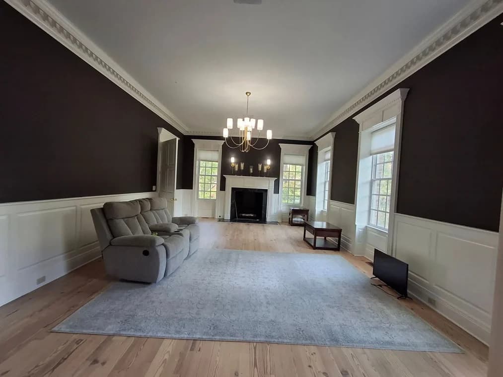 This is an image from Triple G Custom Painting Drywall & Roofing LLC, located in Indiana. The interior photo shows a spacious living room with dark brown walls, white wainscoting, and a white ceiling, freshly painted for a clean and elegant finish.