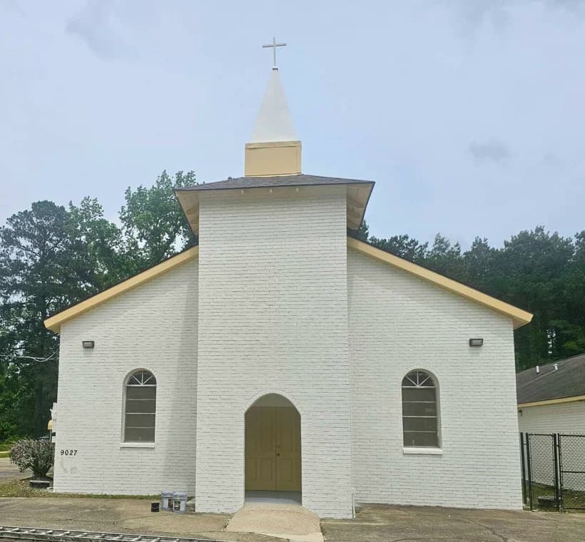 This is an image from LA Painting, located in Louisiana. The image shows a freshly painted white brick church with cream trim, a tall steeple topped with a cross, and arched windows, surrounded by trees under an overcast sky.
