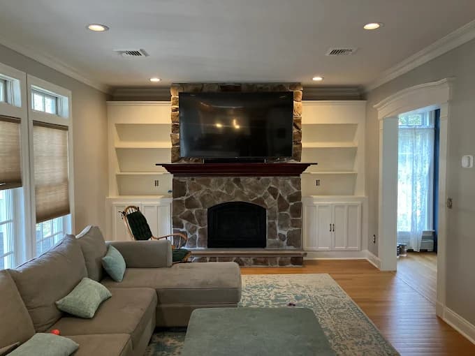 This is an image from Anthony's Painting & Decorating, Inc., located in New York. The photo shows a living room with freshly painted built-in shelves and cabinets framing a stone fireplace beneath a mounted television.