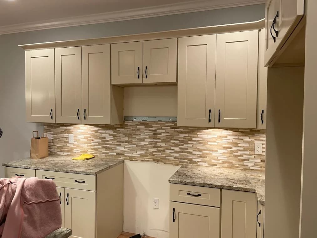 This is an image from Shabby Brown Restore, located in New York. The kitchen shows cream-colored cabinets with black hardware and a stone backsplash illuminated by under-cabinet lighting.
