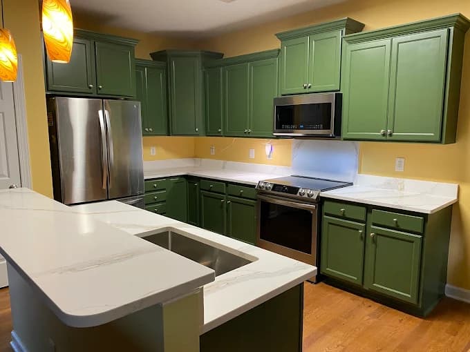 This is an image from Gonzalez Painters & Contractors, Inc., located in North Carolina. The photo shows a modern kitchen with freshly painted green cabinets, yellow walls, stainless steel appliances, and white marble countertops.