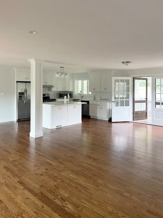 This is an image from CertaPro Painters of Orange County NY, located in New York. The photo shows a bright open-concept kitchen and living area with newly finished hardwood floors, white cabinets, stainless steel appliances, and glass-paneled doors leading to an enclosed porch.