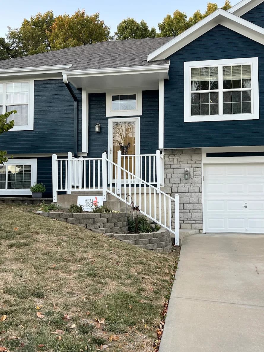 This is an image from Odyssey Painting. The image shows a dark blue two-story home with white trim, a stone accent base, and a front entry stairway leading to a glass storm door.