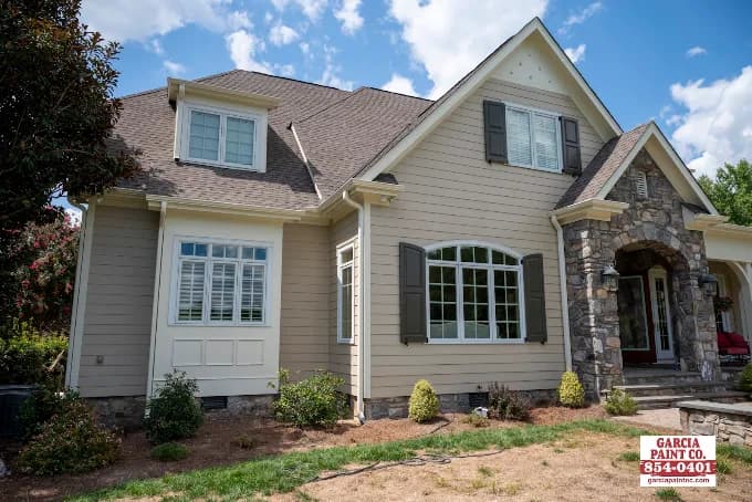 Exterior painting project by Garcia Paint Co in Greensboro, NC. The image shows a beige home with white trim, black shutters, stone entry, and a yard sign.
