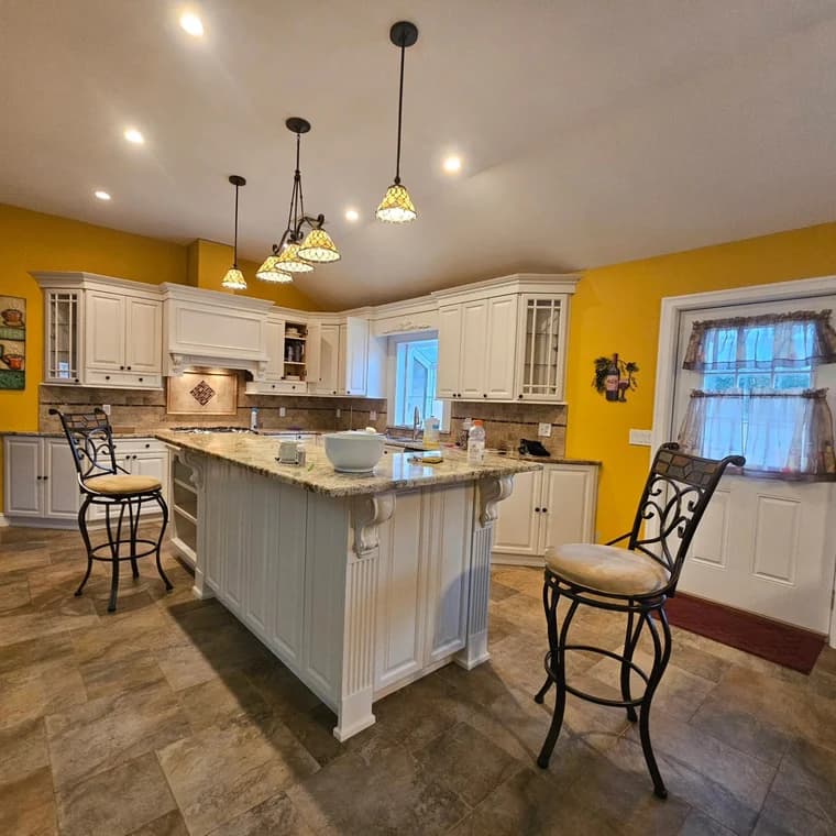 This is an image from JRT Pro Painters, located in New Jersey. The image shows a freshly painted kitchen interior with white cabinets, granite countertops, bright yellow accent walls, and updated lighting fixtures.
