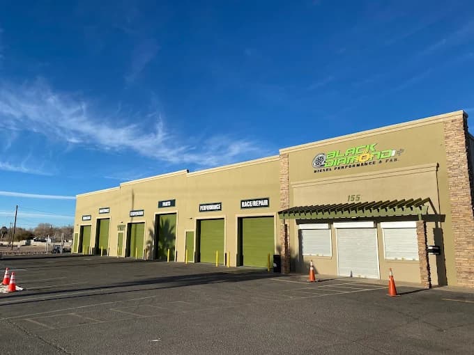 This is an image from Innovative Coatings LLC, located in New Mexico. The photo shows a commercial building with tan walls and green garage doors under clear blue skies, featuring multiple service bays labeled for performance, parts, and race repair.