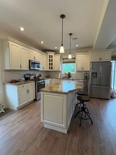 This is an image from Premier Painting Pros, located in New York. The photo shows a spacious kitchen with cream-colored cabinets, granite countertops, stainless steel appliances, pendant lighting, and wood-look flooring.