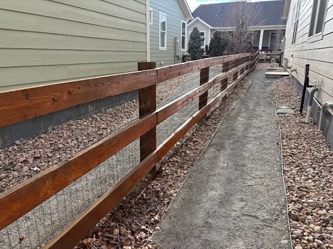 This is an image from Neighborly Fence Staining, LLC company, located in Arvada, CO The image is a freshly stained horizontal split-rail fence with welded wire mesh along a gravel pathway between two suburban homes, completed by Neighborly Fence Staining