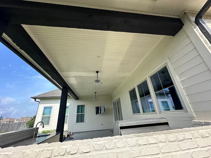 This is an image from Ozuna’s Painting Inc., located in Louisiana. The photo shows a covered patio area with freshly painted white siding, beadboard ceiling, and contrasting black beams, showcasing detailed exterior paintwork and clean finishes.