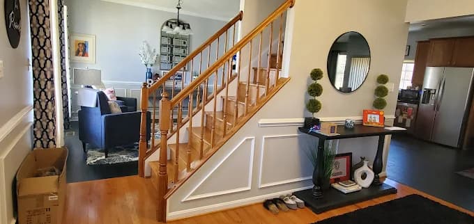 This is an image from CertaPro Painters of Southern Maryland, located in Maryland. The photo shows a freshly painted entryway and staircase with white wainscoting, light gray walls, and natural wood railings leading into a modern living room.