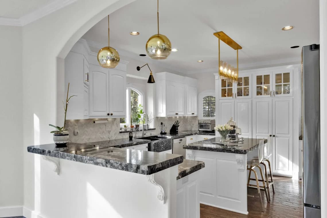 This is an image from Impressions Painting, located in Tulsa, Oklahoma. The photo shows a modern white kitchen with marble countertops, gold pendant lighting, and an island with bar seating.